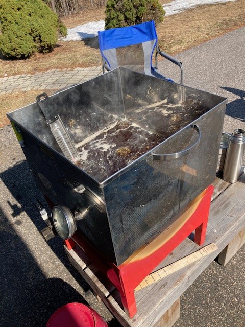 The stainless steel evaporator pan with a thermometer on the side, set outside in the sun after a day's boil, sap residue visible inside.