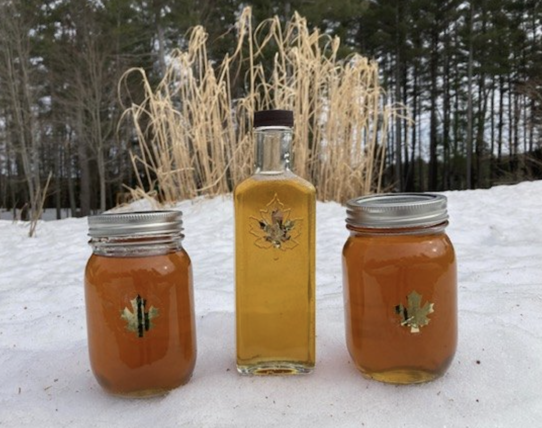 Two mason jars of maple syrup flanking a decorative glass bottle with a gold maple leaf, all standing on snow in front of tall golden grasses.